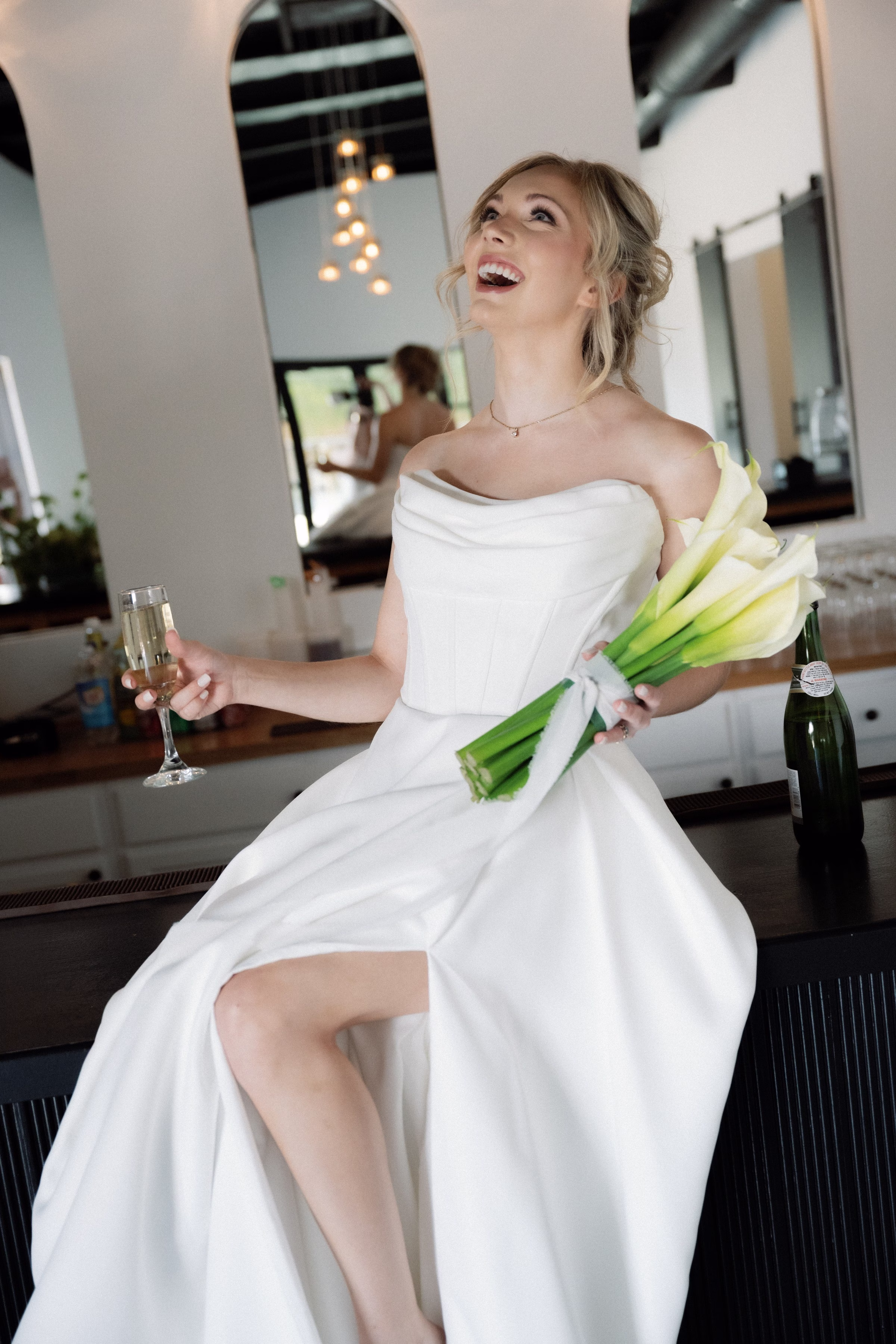 Joyful bride in white gown holding champagne and calla lily bouquet at The Foxwood wedding venue in Hill Country Texas