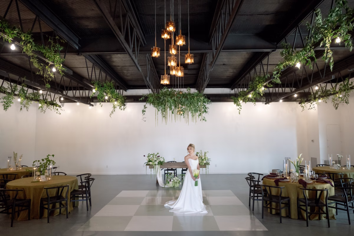 Bride in white gown stands in industrial wedding reception space with exposed beams, hanging greenery, and warm lighting at The Foxwood venue in Hill Country Texas