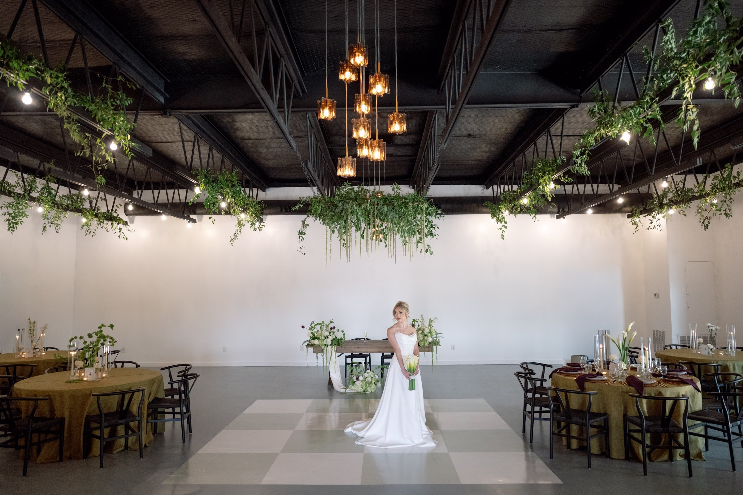 Bride in white gown stands in industrial wedding reception space with exposed beams, hanging greenery, and warm lighting at The Foxwood venue in Hill Country Texas
