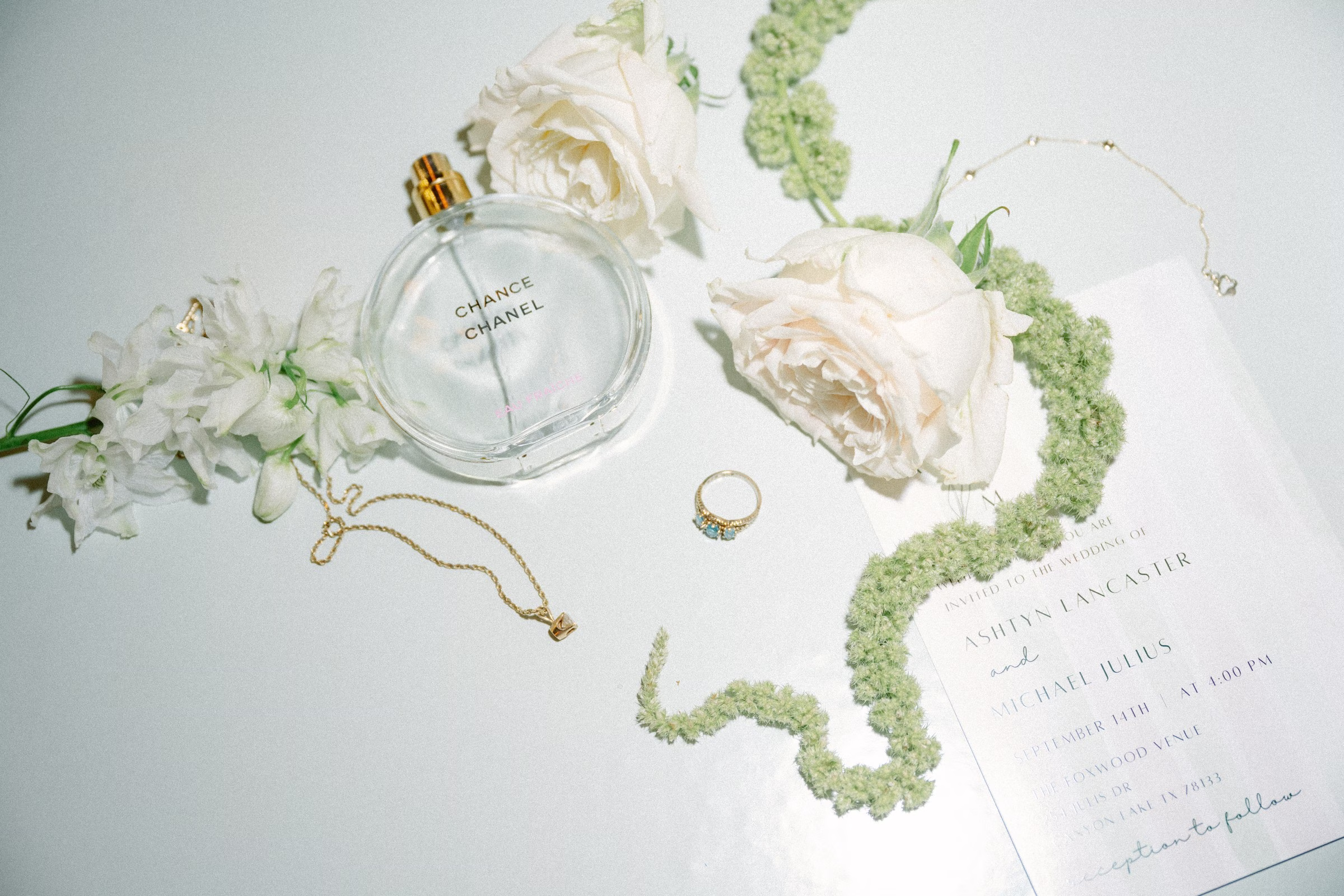 Elegant bridal details flat lay featuring Chanel perfume, jewelry, white roses and wedding invitation at The Foxwood Hill Country Texas wedding venue