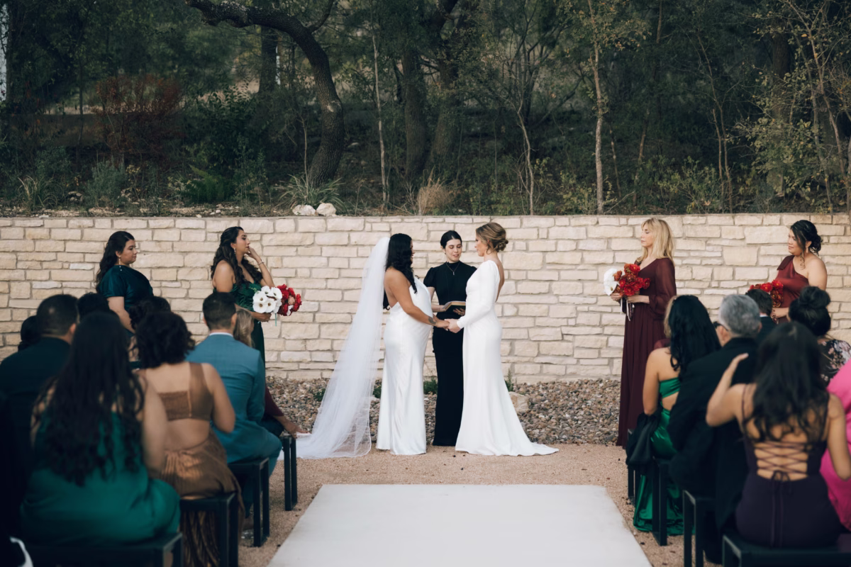 Two brides exchanging vows during outdoor wedding ceremony at The Foxwood Hill Country Texas venue with bridesmaids holding red bouquets