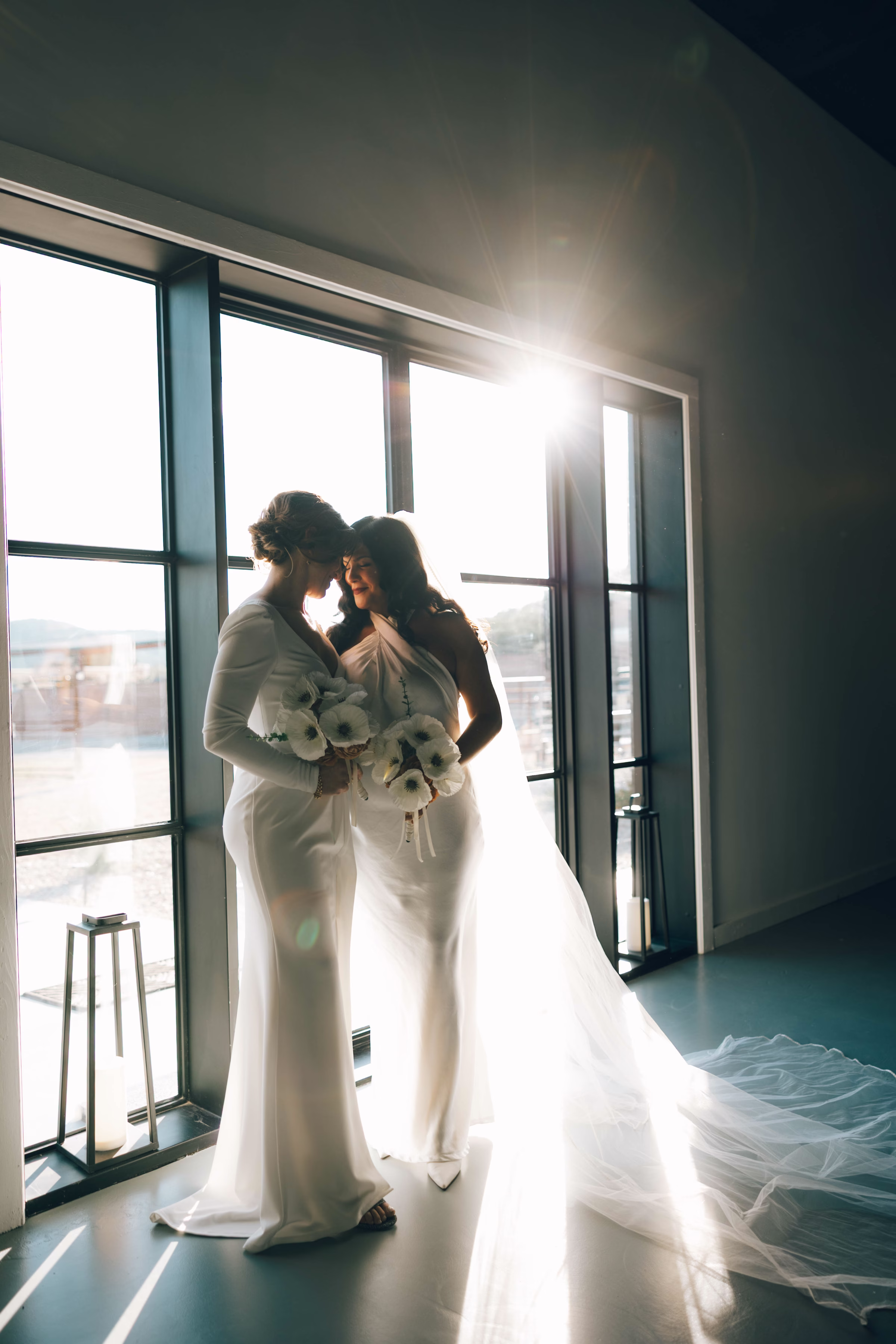Two brides in white dresses sharing an intimate moment by large windows at The Foxwood wedding venue in Hill Country Texas