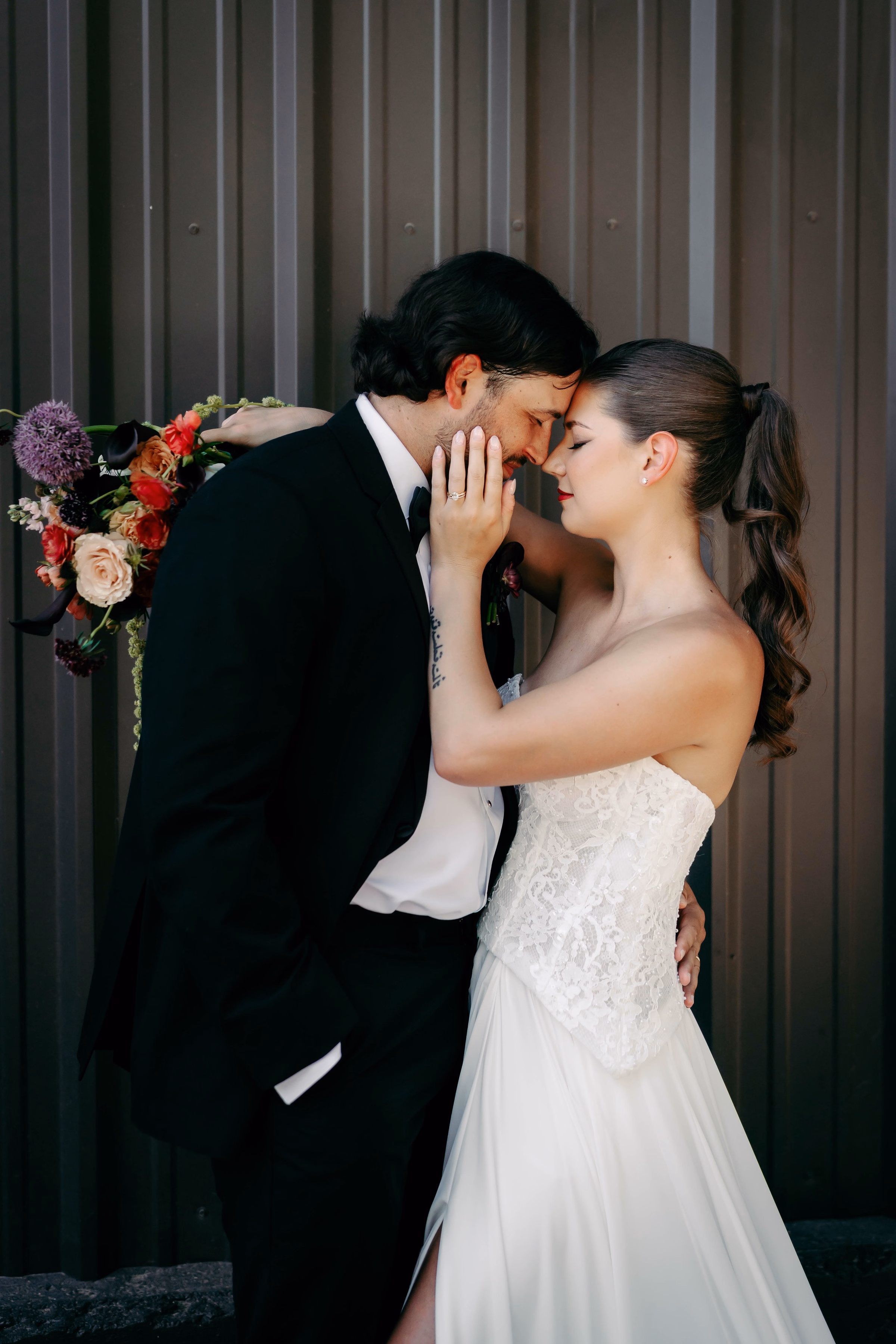 Bride in strapless lace gown and groom in black tuxedo share an intimate forehead touch moment at The Foxwood wedding venue in the Texas Hill Country, with a vibrant bridal bouquet featuring red coral and purple blooms.