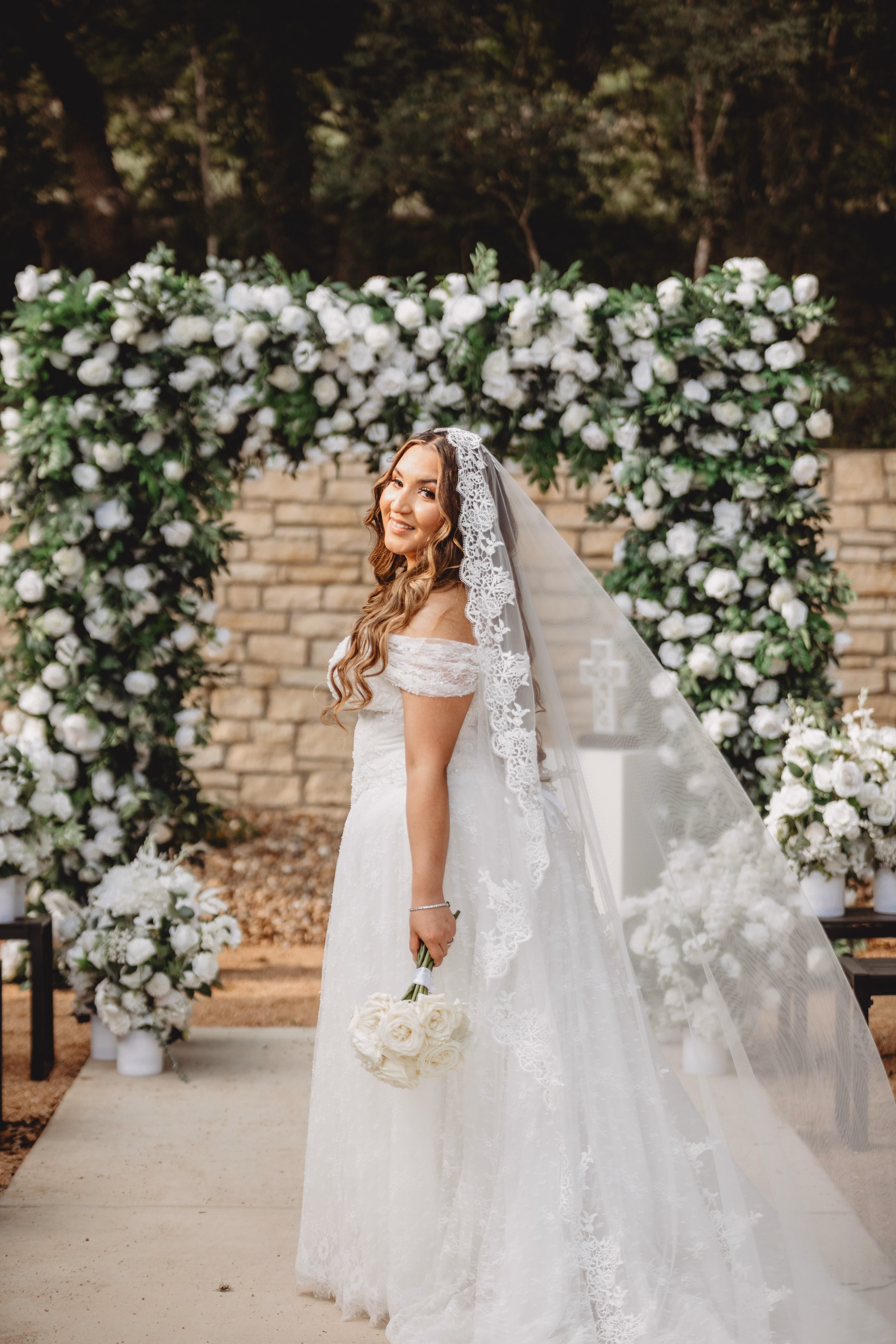 Bride in off-the-shoulder lace gown and cathedral veil holding white rose bouquet in front of a lush white floral arch at The Foxwood wedding venue in the Texas Hill Country.