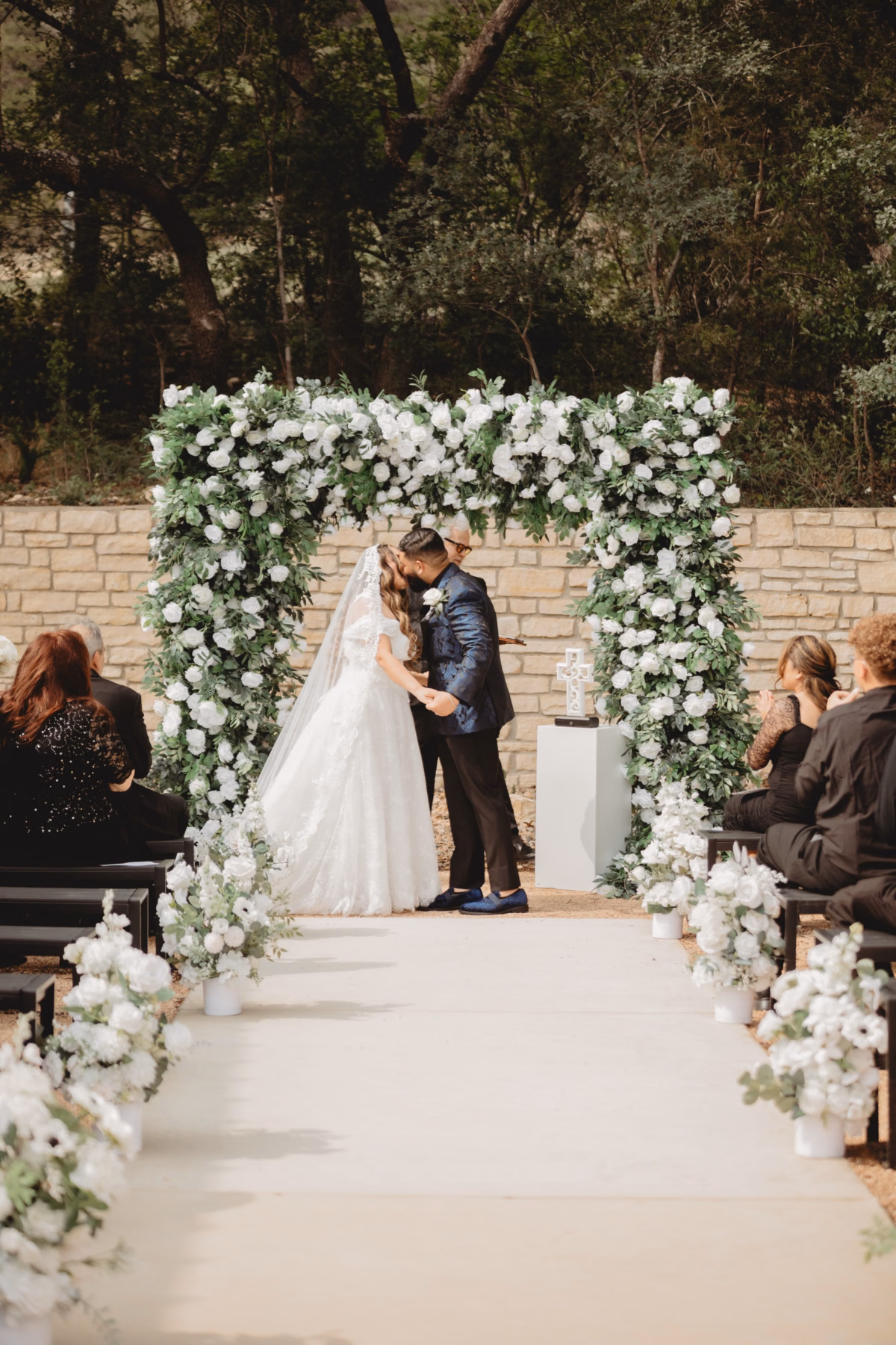 Bride and groom share their first kiss under a lush white floral arch at The Foxwood wedding venue in the Texas Hill Country.