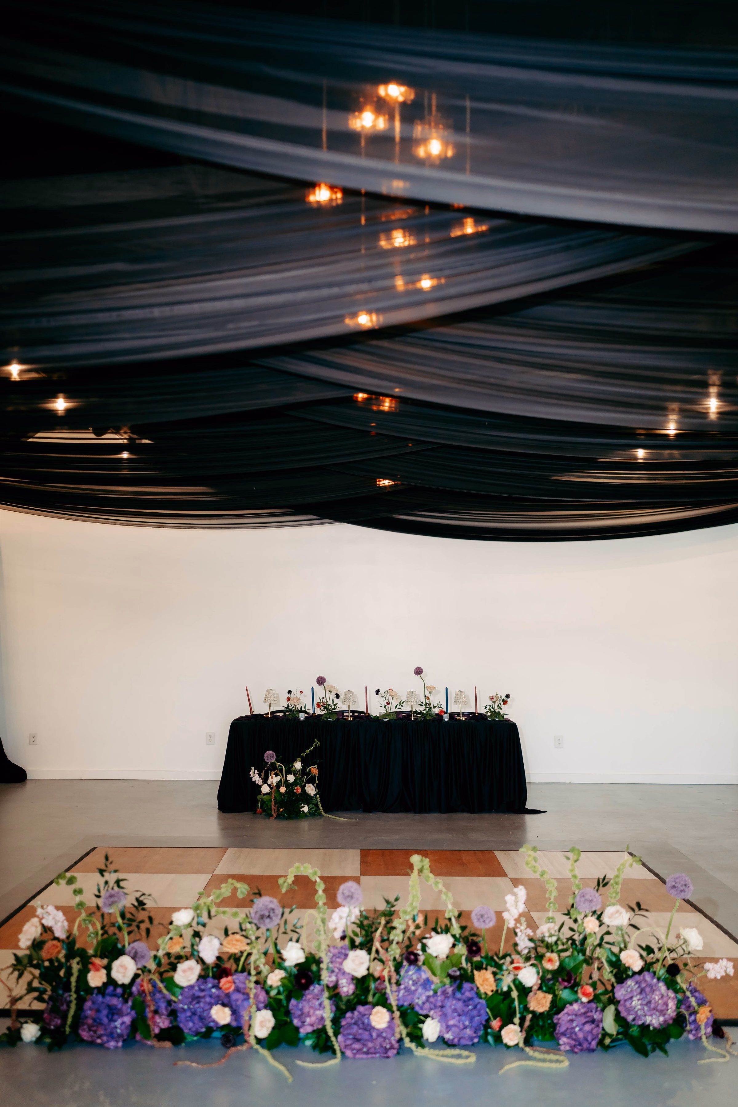 Dramatic sweetheart table with dark linen, candles, and wildflower arrangements set against a white wall beneath sweeping dark ceiling draping, with a lush purple hydrangea and white rose floor floral installation on a checkered dance floor at The Foxwood Hill Country Texas wedding venue.