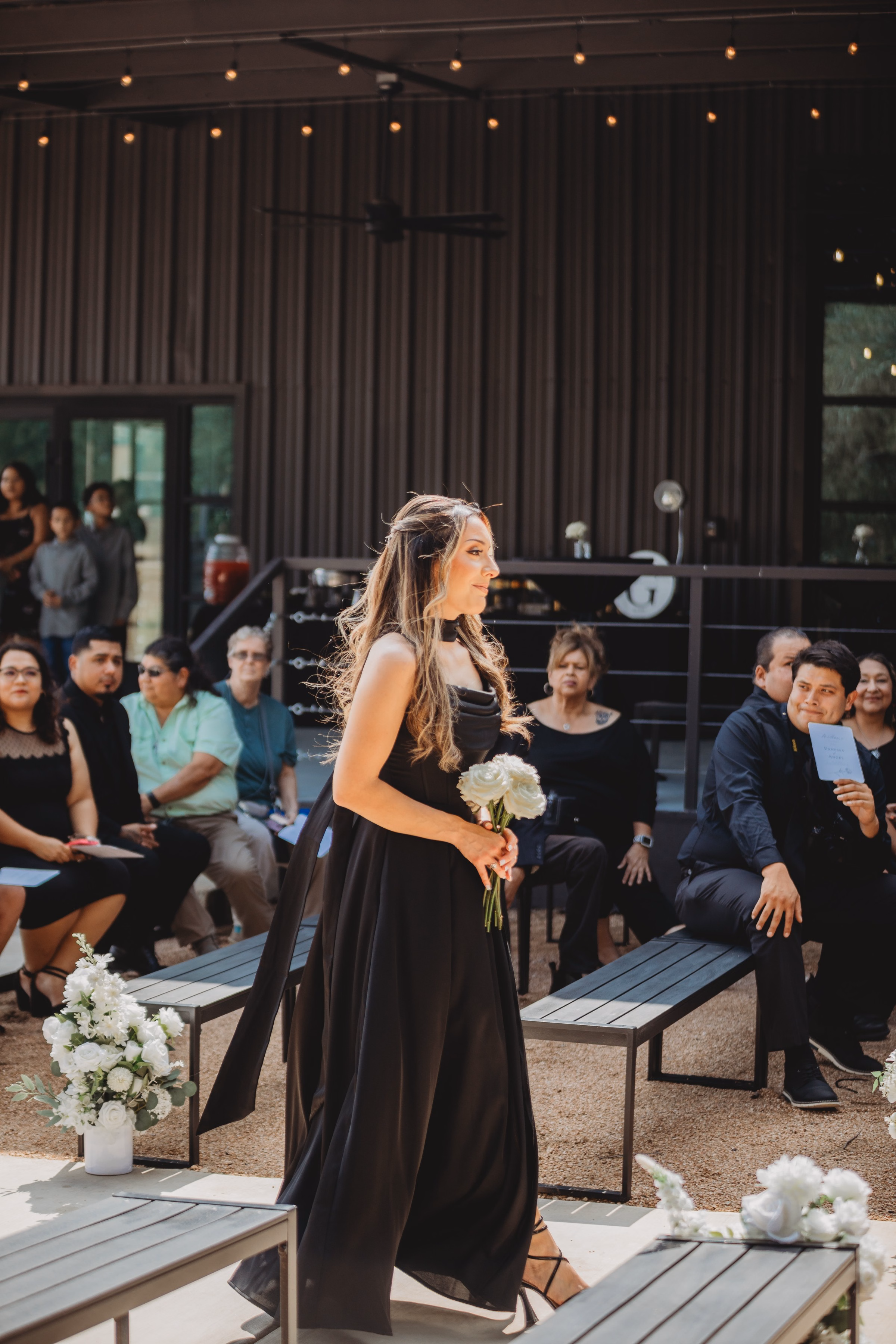 A bridesmaid in a black gown walks the outdoor ceremony aisle holding white roses at The Foxwood wedding venue in the Texas Hill Country.