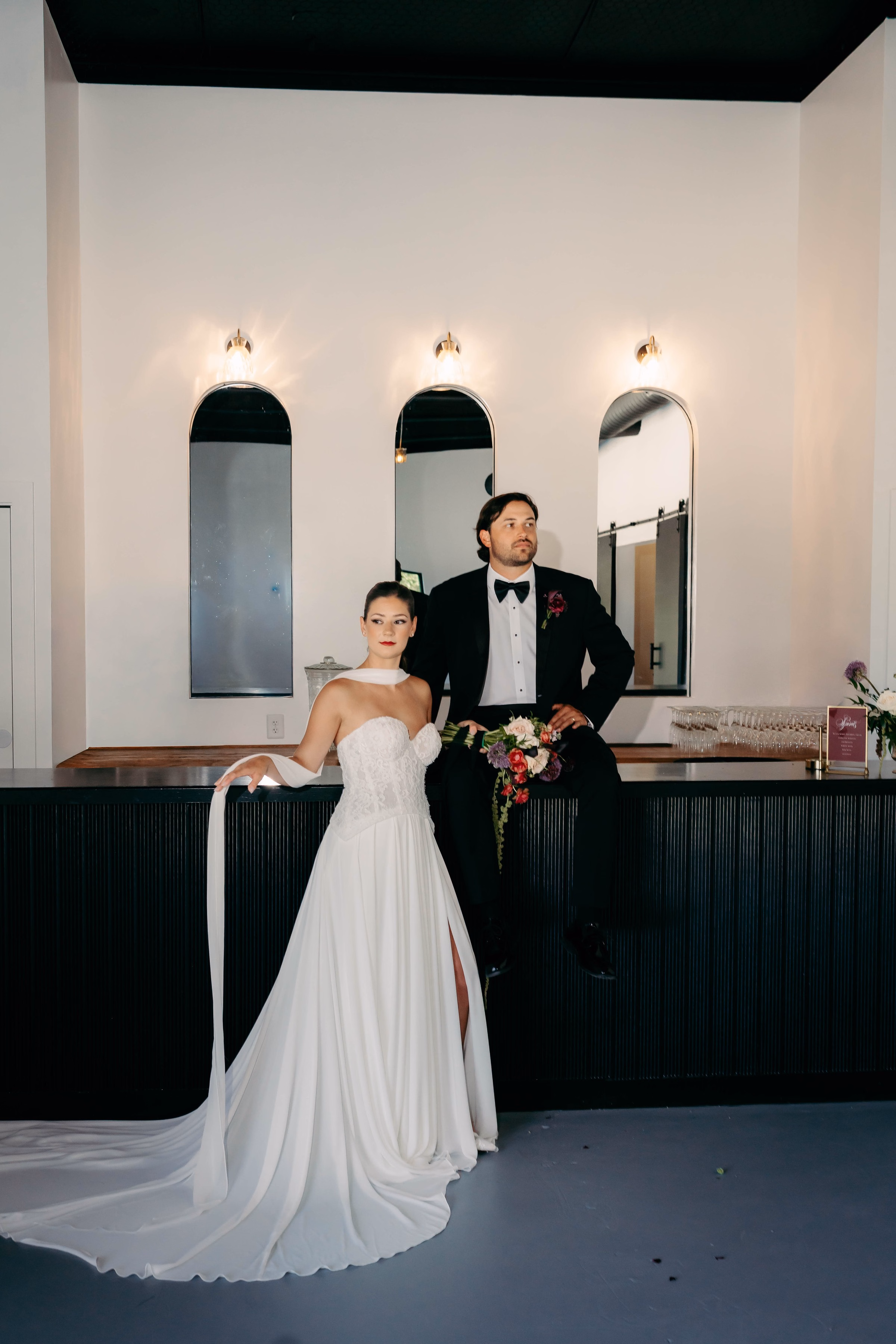 Bride in strapless A-line gown with halter sash and groom in black tuxedo pose at the modern dark bar at The Foxwood wedding venue in Hill Country Texas.