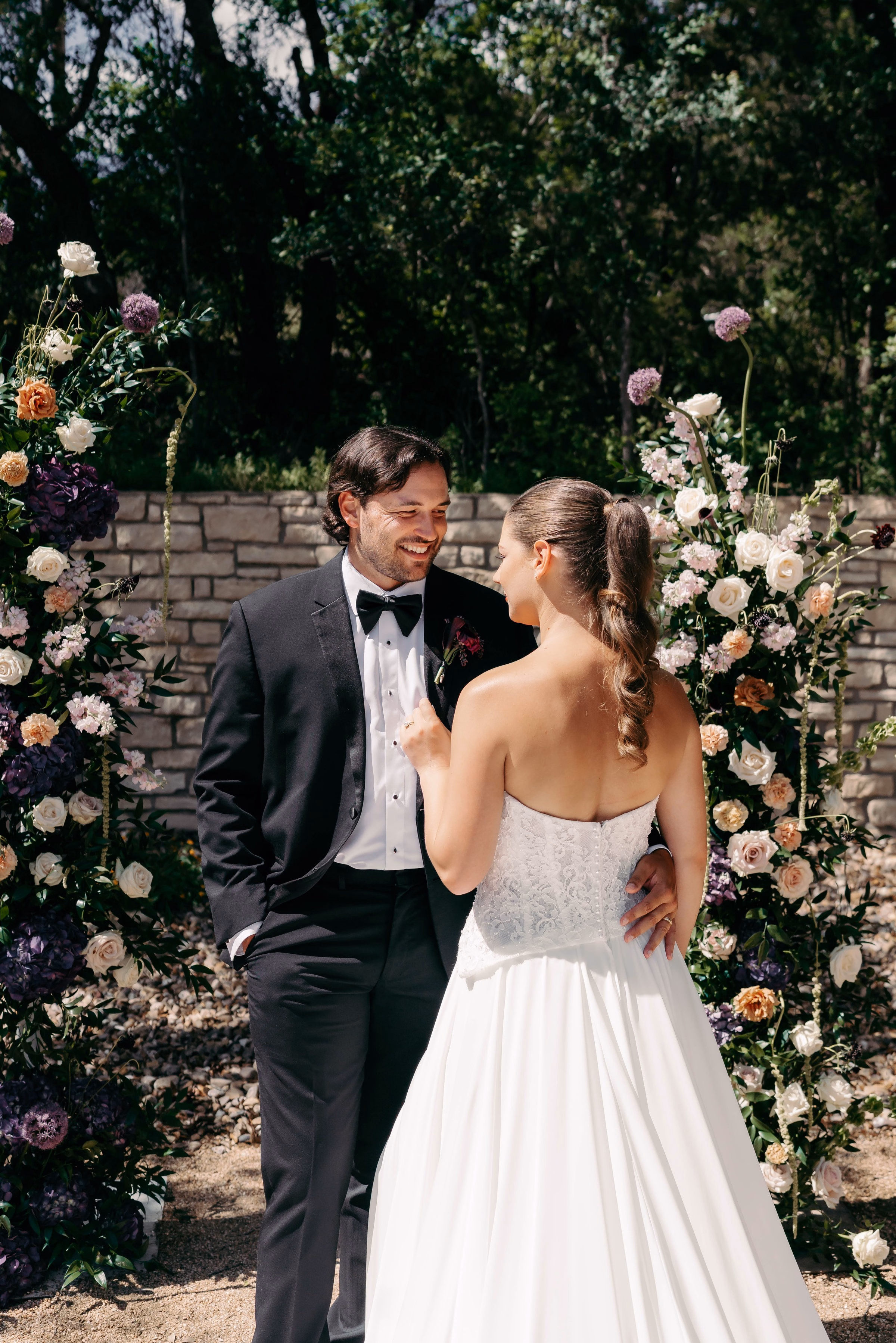 Bride and groom share a joyful first look moment in front of a lush floral arch with purple, blush, and ivory blooms at The Foxwood Hill Country Texas wedding venue.