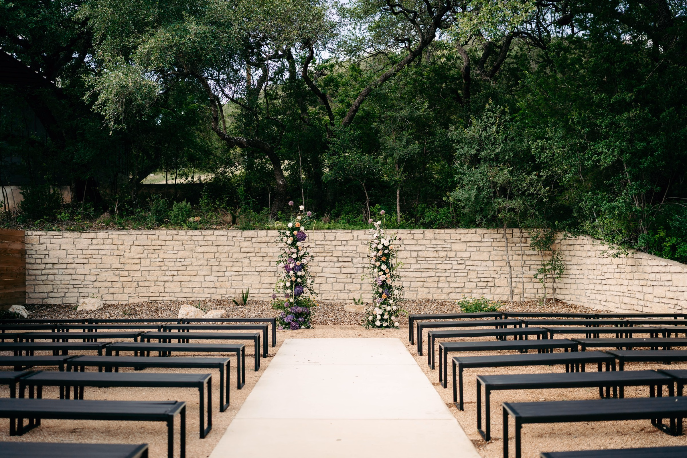 Outdoor wedding ceremony at The Foxwood in the Texas Hill Country featuring black modern benches lining a white aisle runner leading to two floral column arrangements of purple and blush blooms against a limestone wall backdrop.
